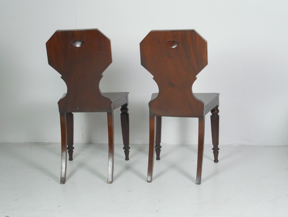 A pair of antique Regency mahogany hall chairs featuring octagonal carved backs with central shield motifs, solid timber seats, and turned front legs. View VI