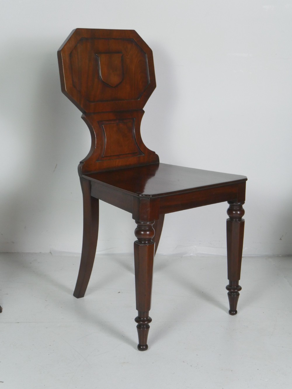 A pair of antique Regency mahogany hall chairs featuring octagonal carved backs with central shield motifs, solid timber seats, and turned front legs. View VIII