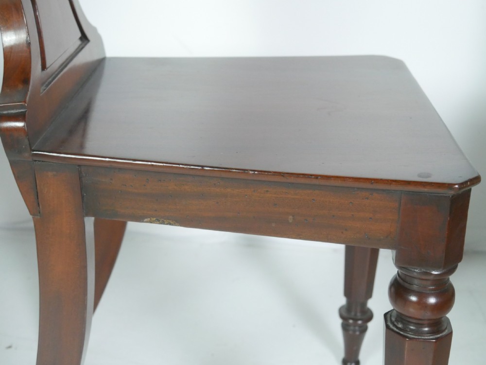 A pair of antique Regency mahogany hall chairs featuring octagonal carved backs with central shield motifs, solid timber seats, and turned front legs. View XIV