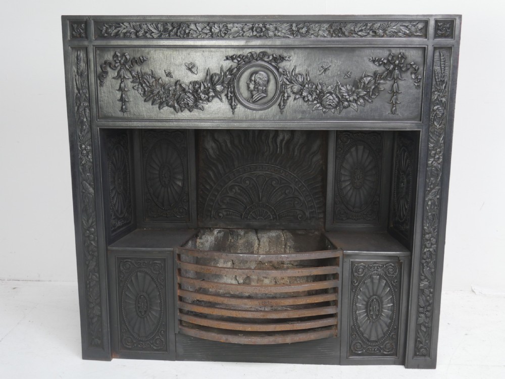 A dark cast iron register grate decorated with carved floral swags, a central profile medallion, and a fan-motif back plate above a curved front grate.