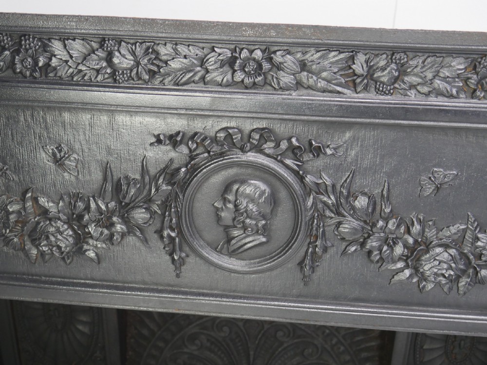 A dark cast iron register grate decorated with carved floral swags, a central profile medallion, and a fan-motif back plate above a curved front grate. View V