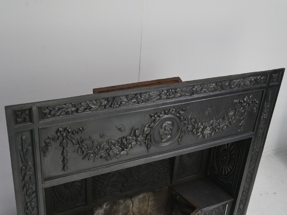 A dark cast iron register grate decorated with carved floral swags, a central profile medallion, and a fan-motif back plate above a curved front grate. View VI