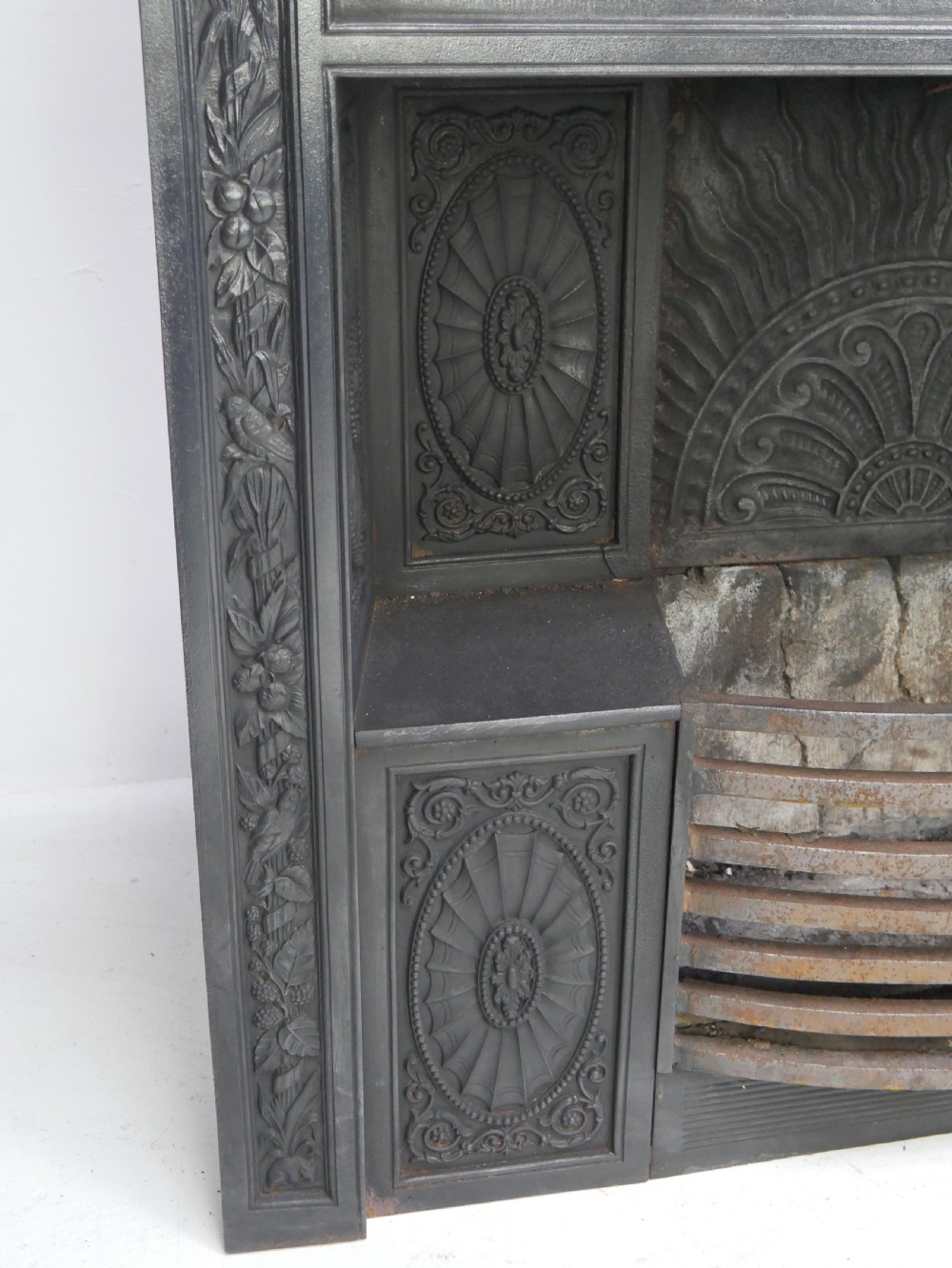 A dark cast iron register grate decorated with carved floral swags, a central profile medallion, and a fan-motif back plate above a curved front grate. View VII