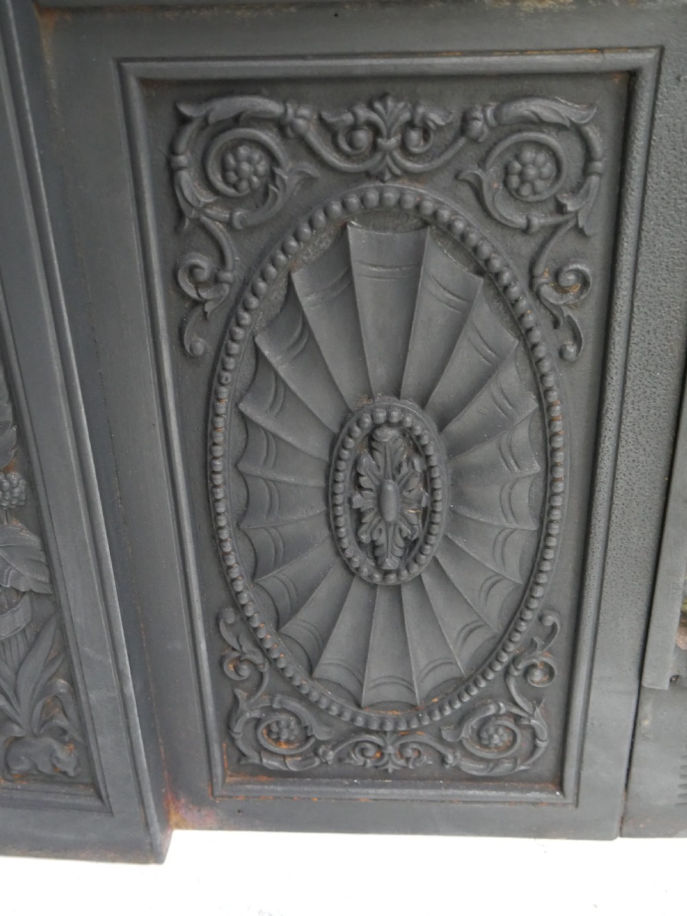 A dark cast iron register grate decorated with carved floral swags, a central profile medallion, and a fan-motif back plate above a curved front grate. View IX