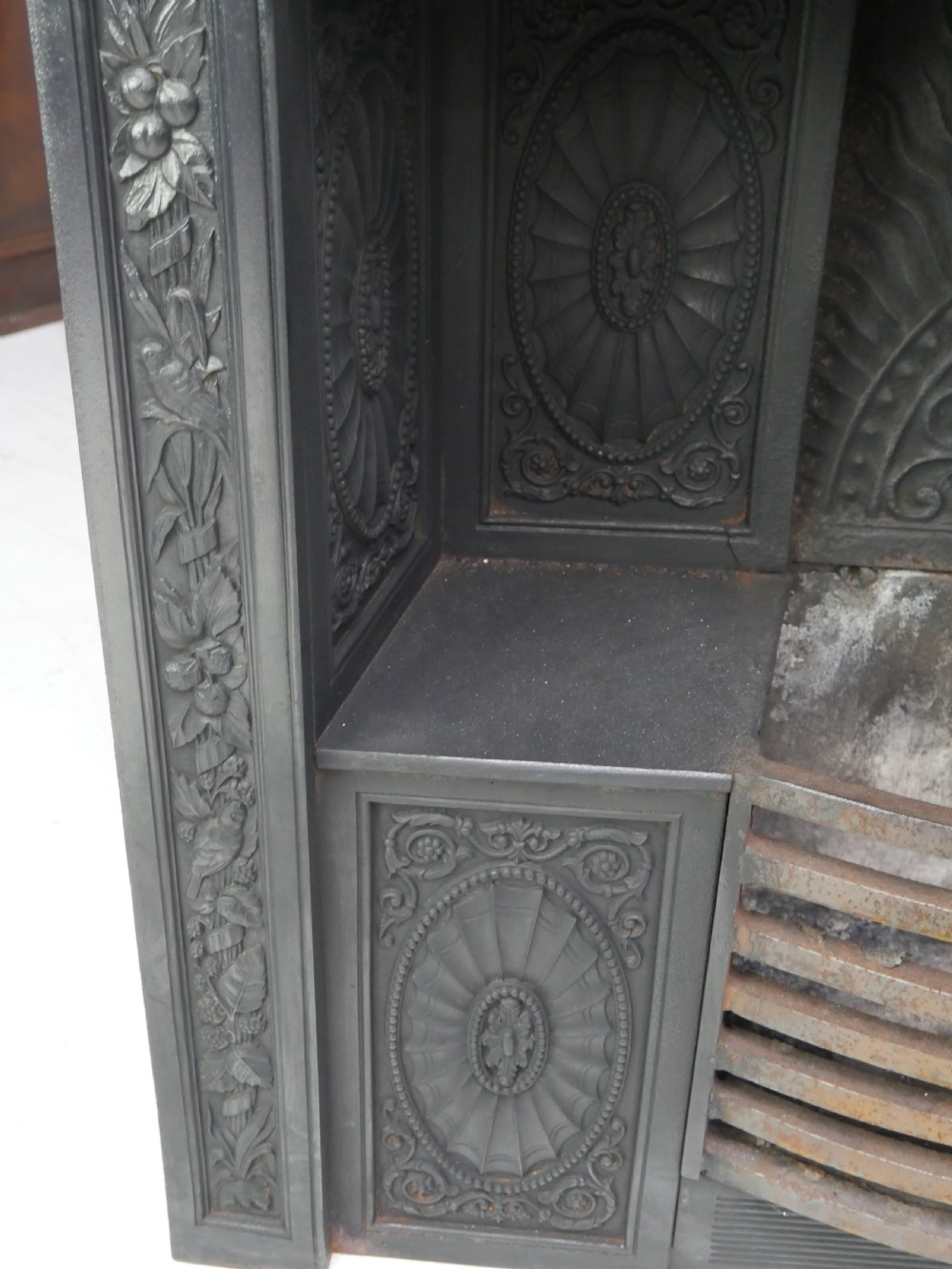 A dark cast iron register grate decorated with carved floral swags, a central profile medallion, and a fan-motif back plate above a curved front grate. View XVIII