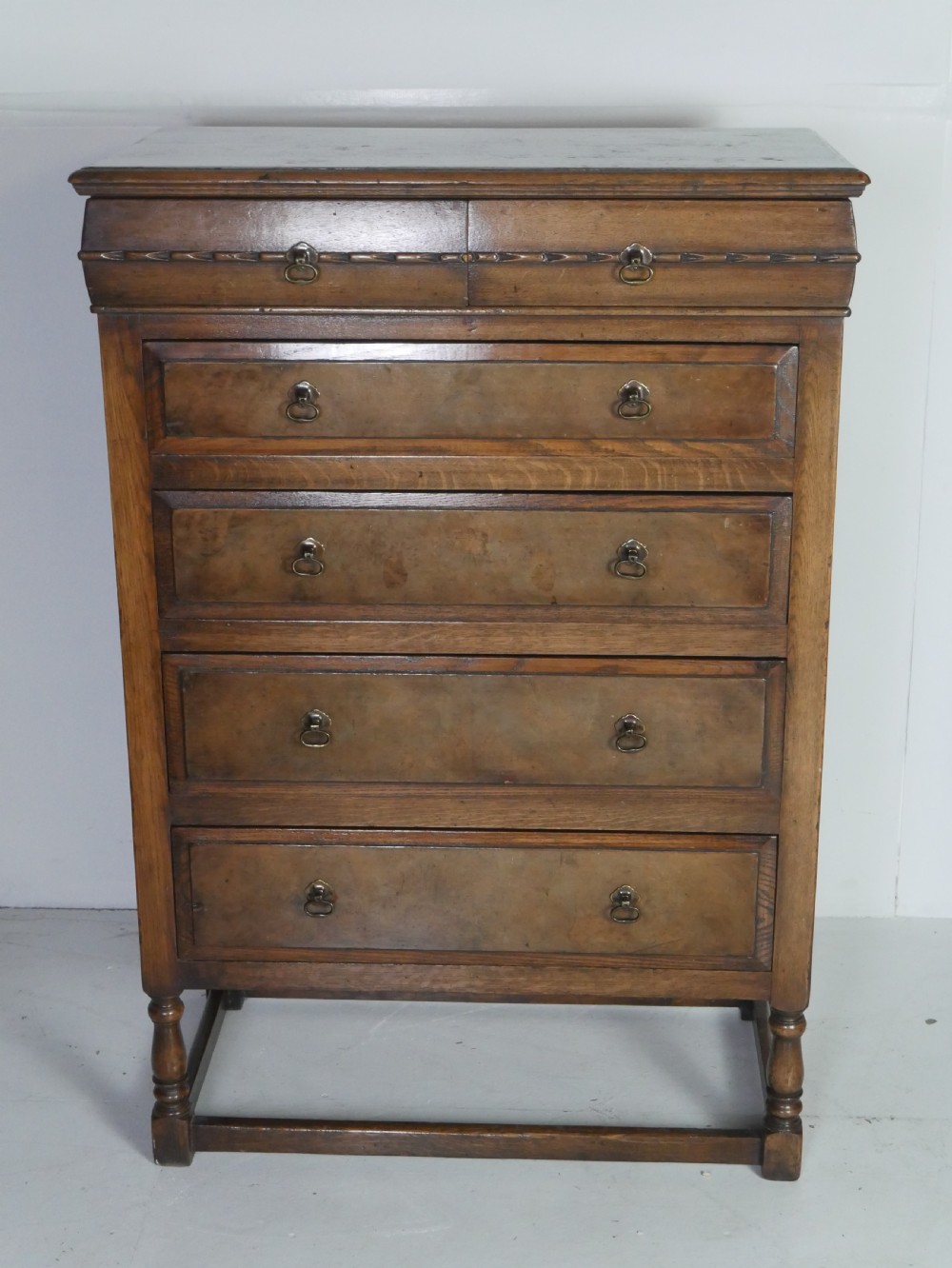 A tall wooden chest of drawers constructed from oak and walnut, featuring five drawers, panelled sides, turned legs with a stretcher base, and brass drop handles. View II
