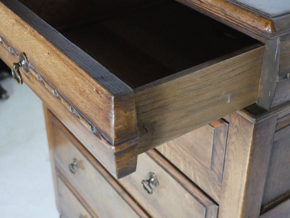A tall wooden chest of drawers constructed from oak and walnut, featuring five drawers, panelled sides, turned legs with a stretcher base, and brass drop handles. View III