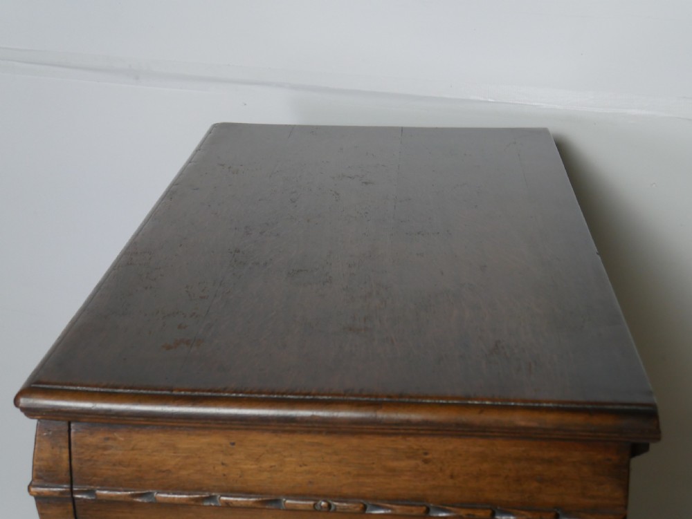 A tall wooden chest of drawers constructed from oak and walnut, featuring five drawers, panelled sides, turned legs with a stretcher base, and brass drop handles. View VI