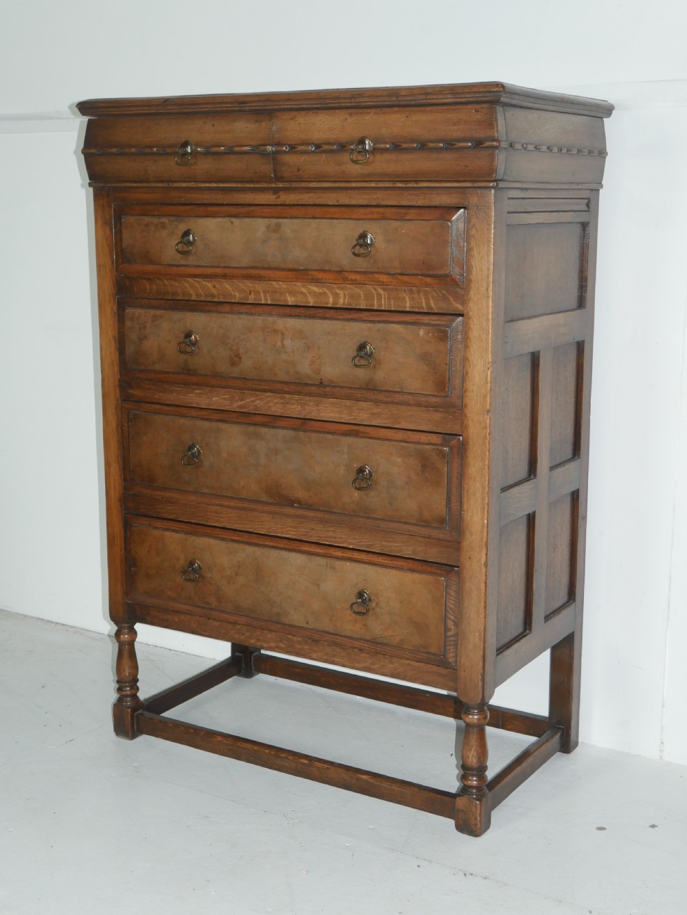 A tall wooden chest of drawers constructed from oak and walnut, featuring five drawers, panelled sides, turned legs with a stretcher base, and brass drop handles. View XI