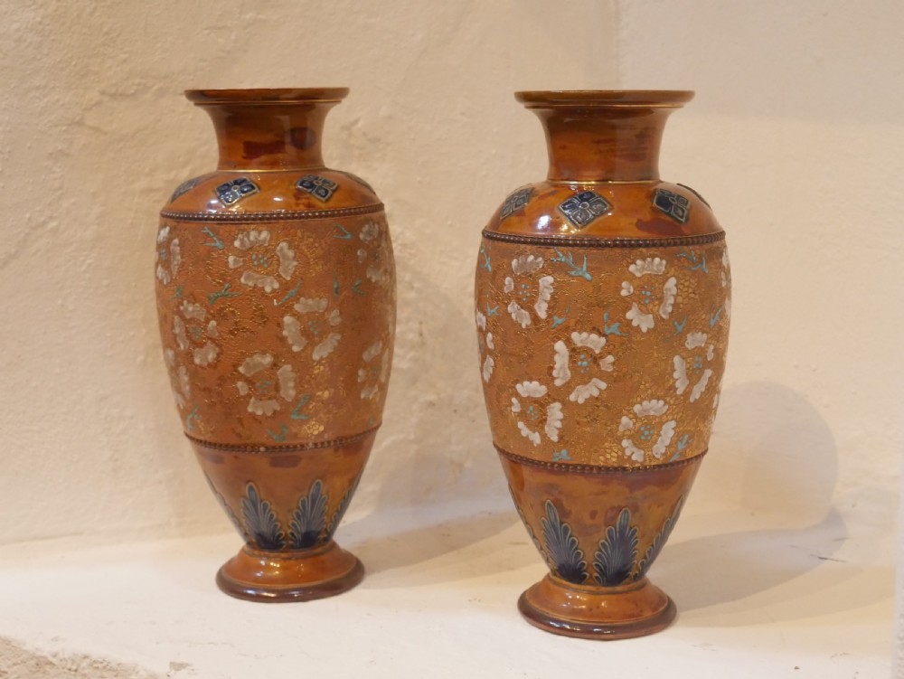 A pair of antique Royal Doulton stoneware vases featuring caramel brown glazing, white floral decorations, and blue foliate accents around the base. View XII