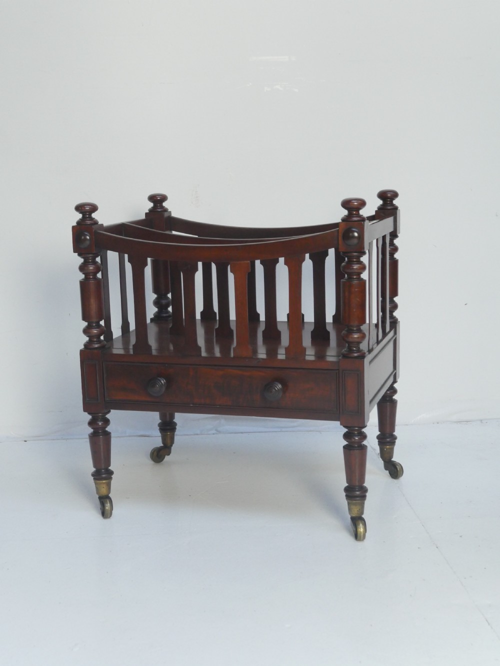 A dark mahogany wooden Canterbury magazine rack featuring slatted dividers, a single drawer with turned knobs, and turned legs with brass castors.