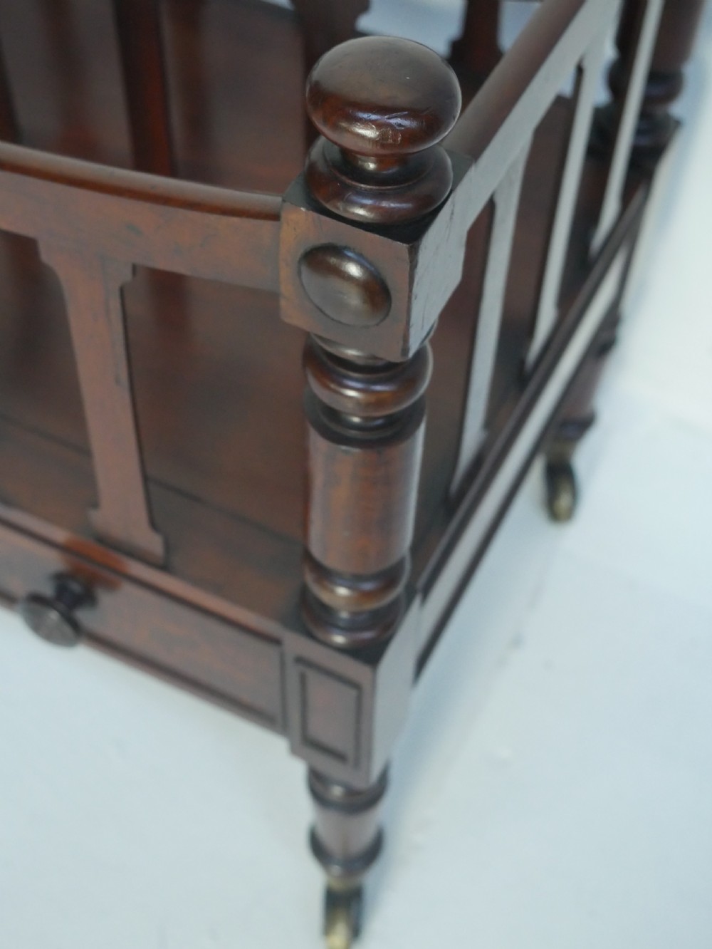 A dark mahogany wooden Canterbury magazine rack featuring slatted dividers, a single drawer with turned knobs, and turned legs with brass castors. View V
