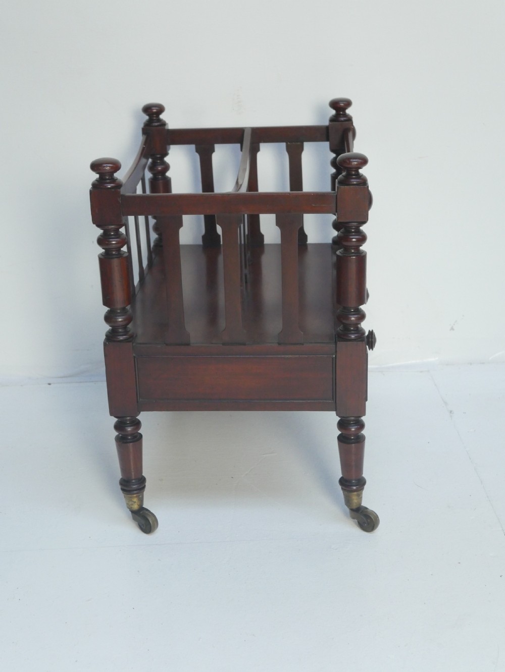 A dark mahogany wooden Canterbury magazine rack featuring slatted dividers, a single drawer with turned knobs, and turned legs with brass castors. View VIII