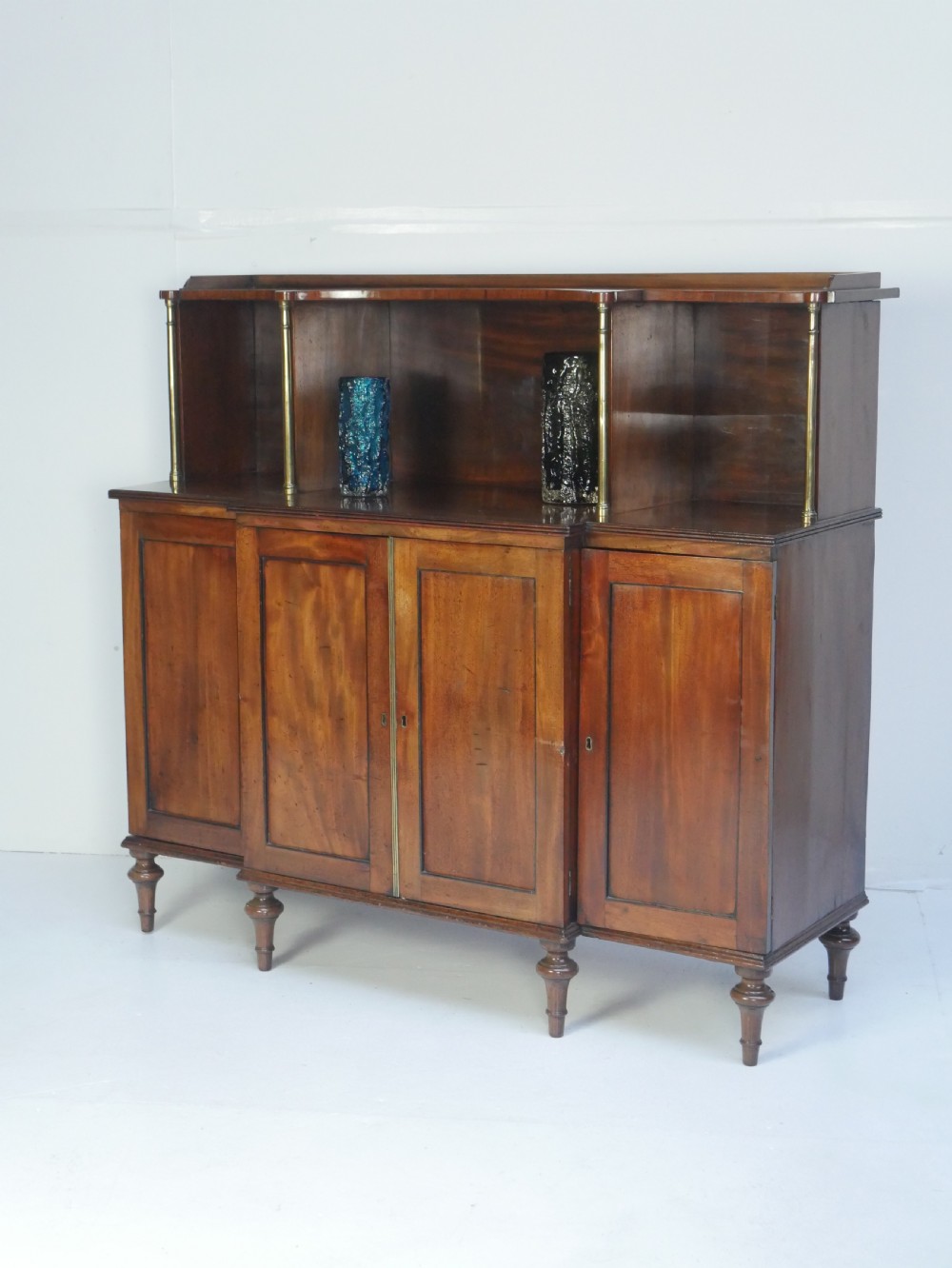 A Georgian mahogany chiffonier featuring an upper display shelf supported by brass columns above panelled cupboard doors and turned legs.