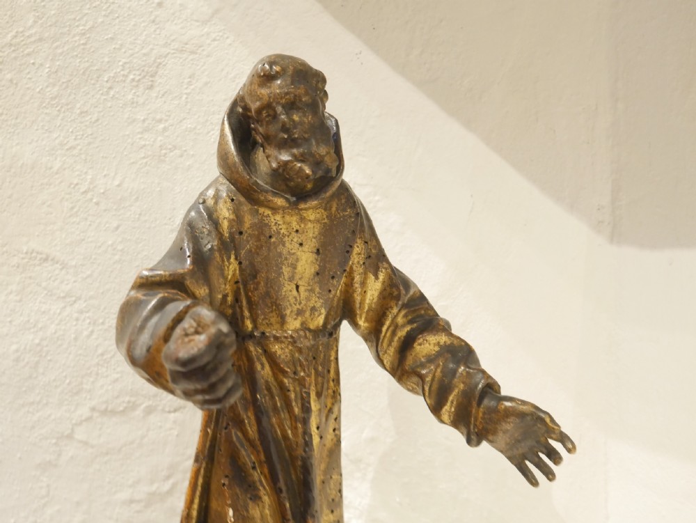 A carved giltwood figure depicting Saint Francis in draped monastic robes standing on a dark wooden plinth. View II
