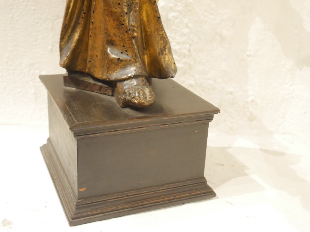 A carved giltwood figure depicting Saint Francis in draped monastic robes standing on a dark wooden plinth. View III