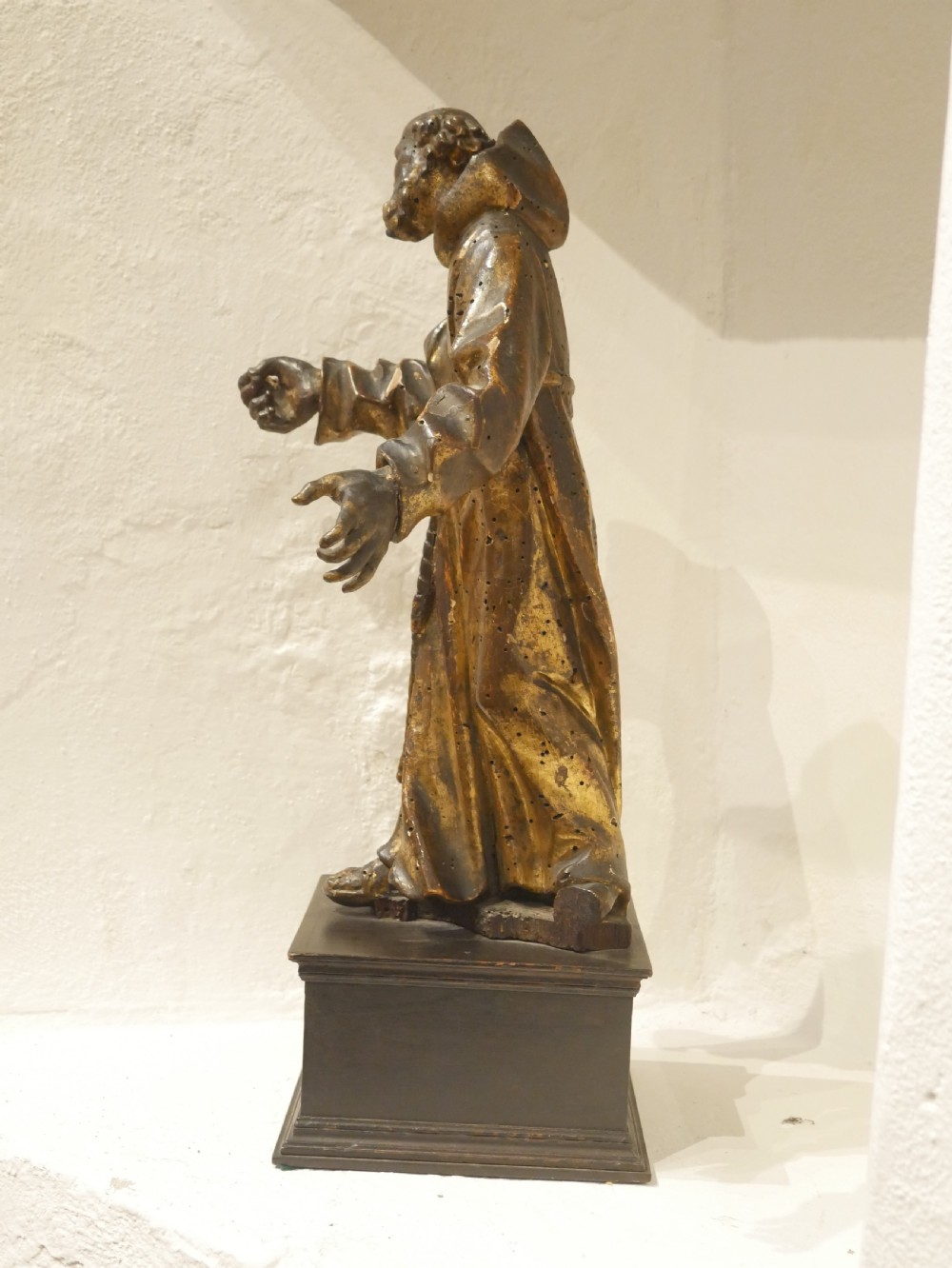 A carved giltwood figure depicting Saint Francis in draped monastic robes standing on a dark wooden plinth. View X