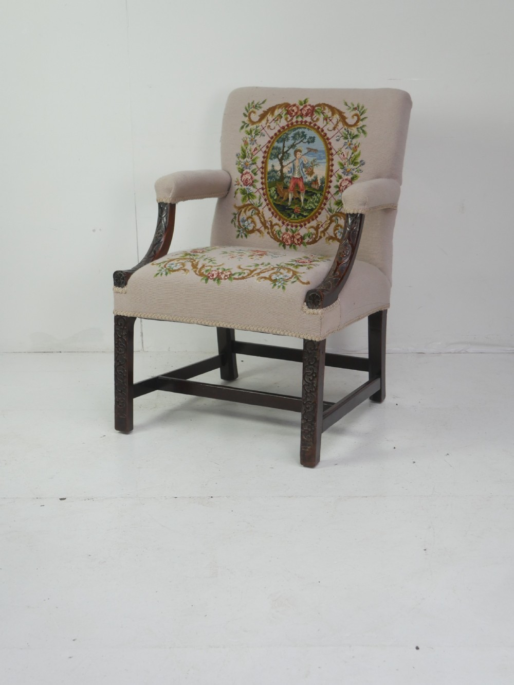 A Georgian Gainsborough armchair with dark carved mahogany legs, upholstered in beige needlepoint tapestry featuring a central pastoral scene and floral borders.