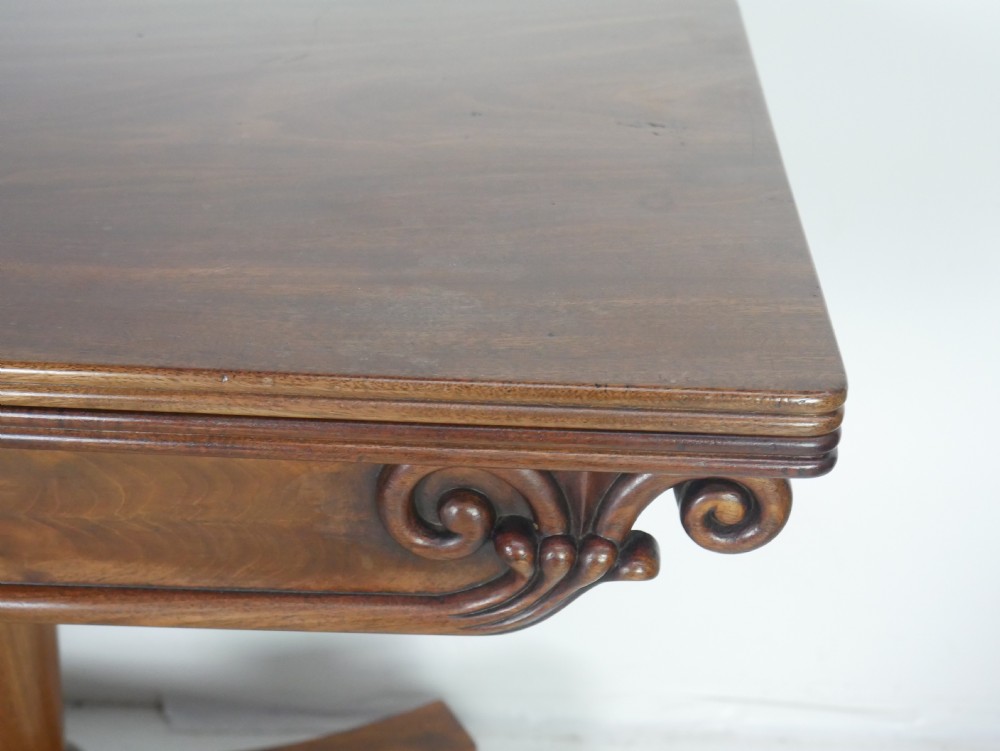 A 19th-century mahogany turn-over table featuring a fold-over top, carved corner scroll accents, a tapered pedestal, and a quadform platform base with scroll feet. View VII