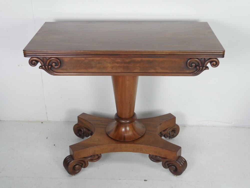 A 19th-century mahogany turn-over table featuring a fold-over top, carved corner scroll accents, a tapered pedestal, and a quadform platform base with scroll feet. View XV
