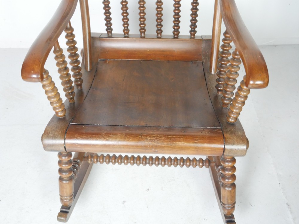 An antique warm brown wooden rocking chair featuring intricate bobbin-turned spindles along the backrest, armrests, legs, and stretchers. View VII
