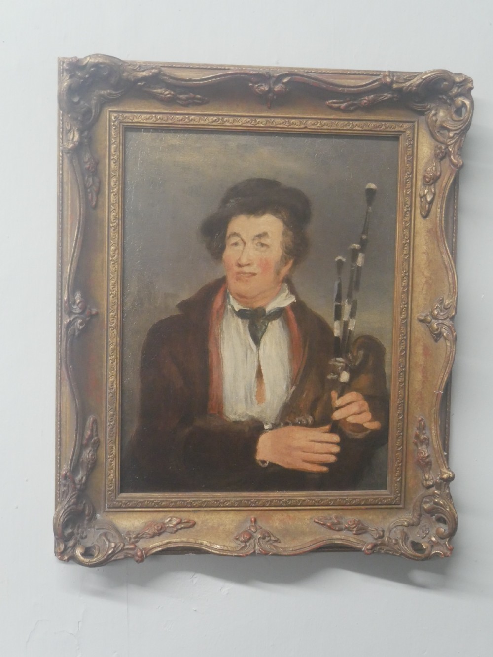 An oil portrait on canvas depicting a male Northumbrian piper holding smallpipes, housed in an ornate carved gilt frame. View VII