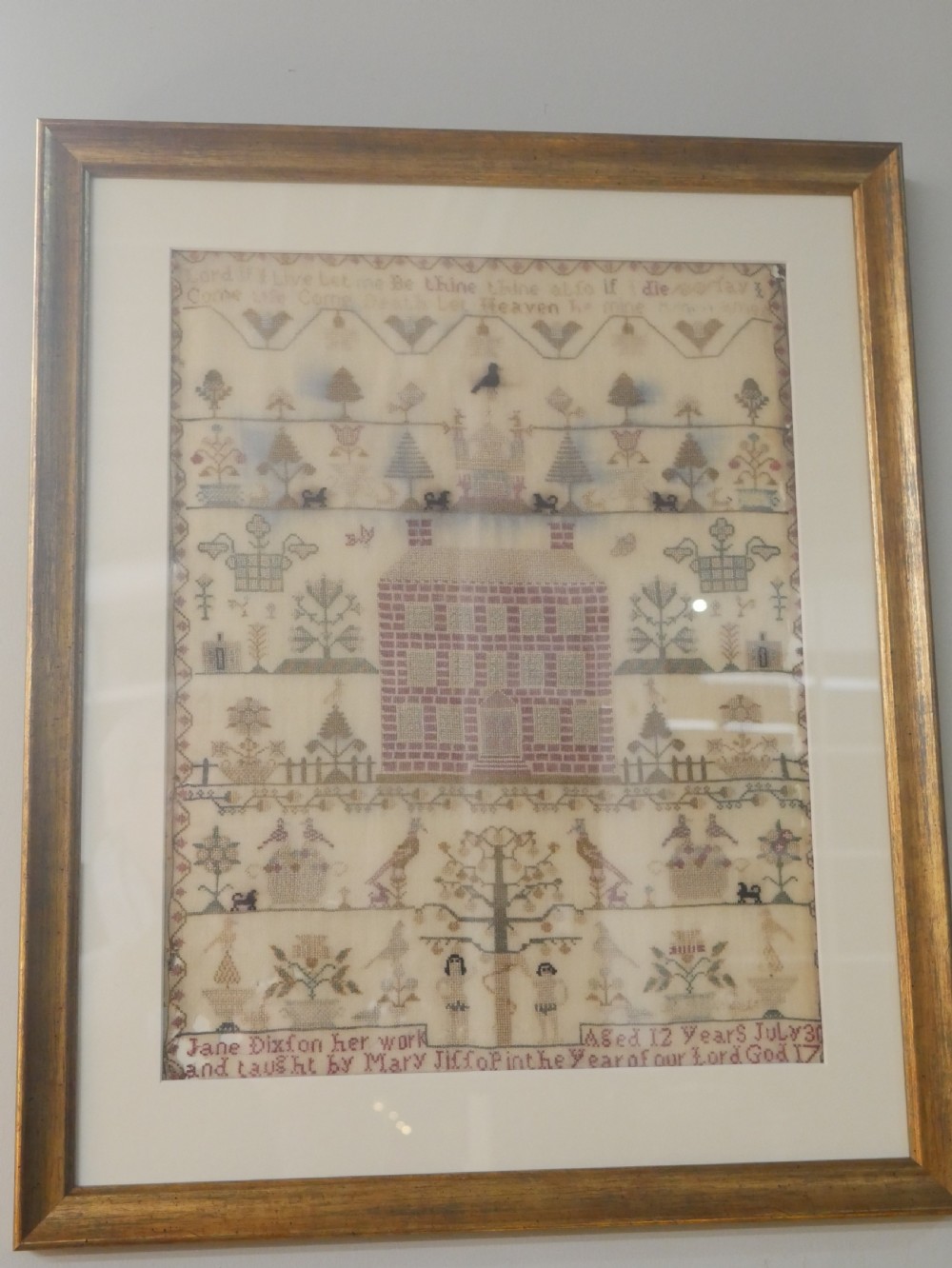 An 18th-century framed needlework sampler featuring a central brick house motif surrounded by trees, animals, figures, and stitched text on a linen ground.