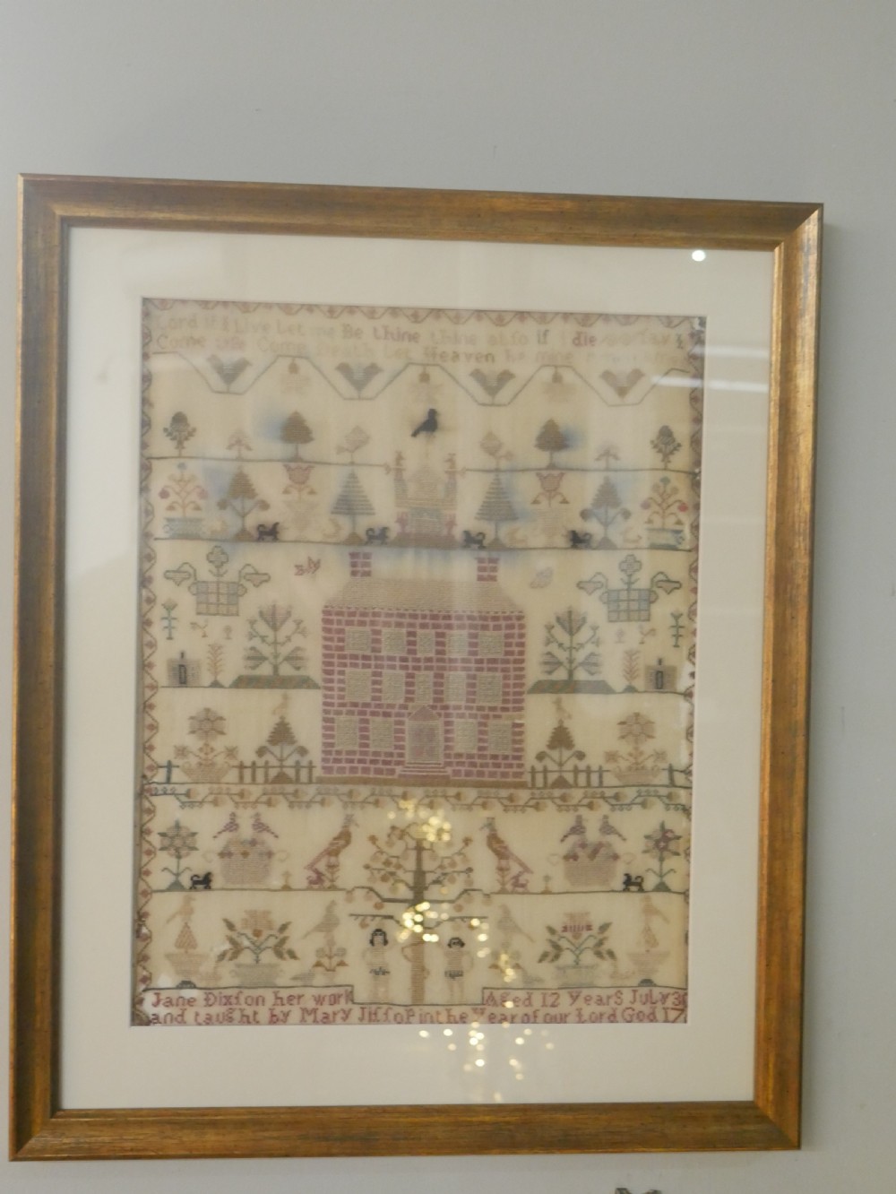 An 18th-century framed needlework sampler featuring a central brick house motif surrounded by trees, animals, figures, and stitched text on a linen ground. View XI