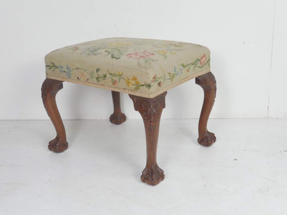 A square upholstered footstool featuring floral needlepoint fabric, carved cabriole legs, and claw and ball feet.