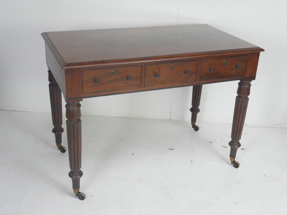 An English Regency mahogany freestanding writing desk featuring three frieze drawers with small turned knobs, supported by four reeded legs on brass castors.