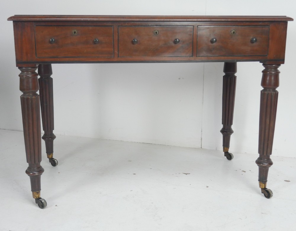 An English Regency mahogany freestanding writing desk featuring three frieze drawers with small turned knobs, supported by four reeded legs on brass castors. View II