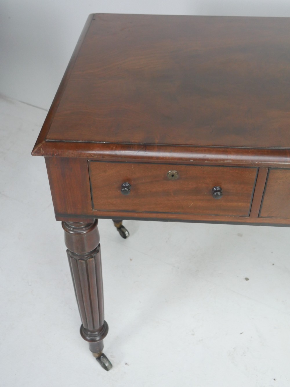 An English Regency mahogany freestanding writing desk featuring three frieze drawers with small turned knobs, supported by four reeded legs on brass castors. View IV