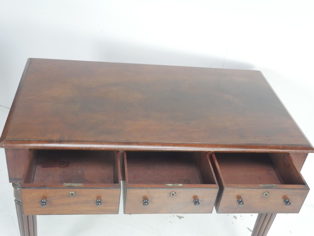 An English Regency mahogany freestanding writing desk featuring three frieze drawers with small turned knobs, supported by four reeded legs on brass castors. View V