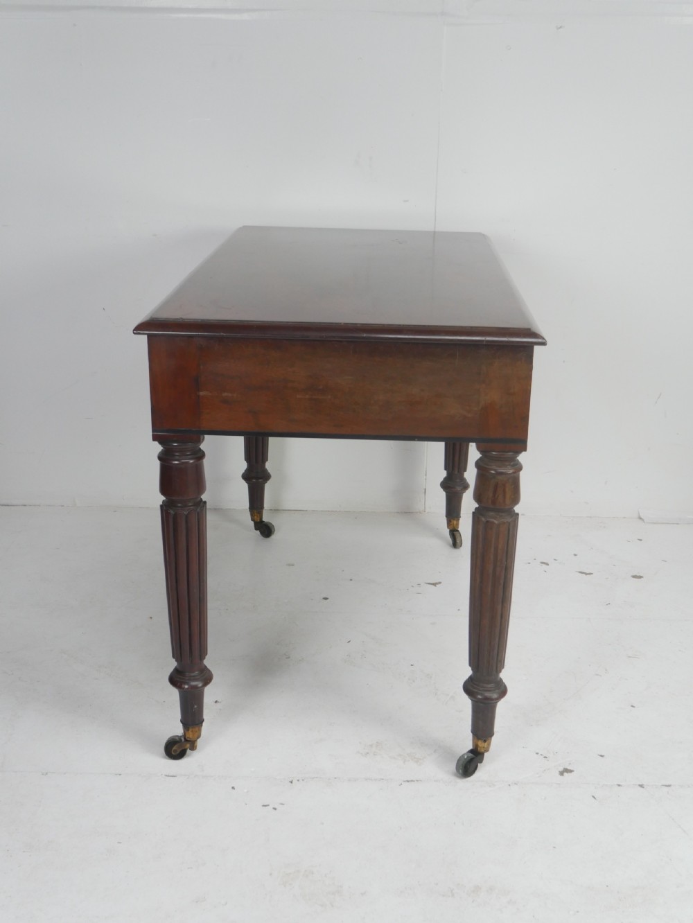 An English Regency mahogany freestanding writing desk featuring three frieze drawers with small turned knobs, supported by four reeded legs on brass castors. View VIII