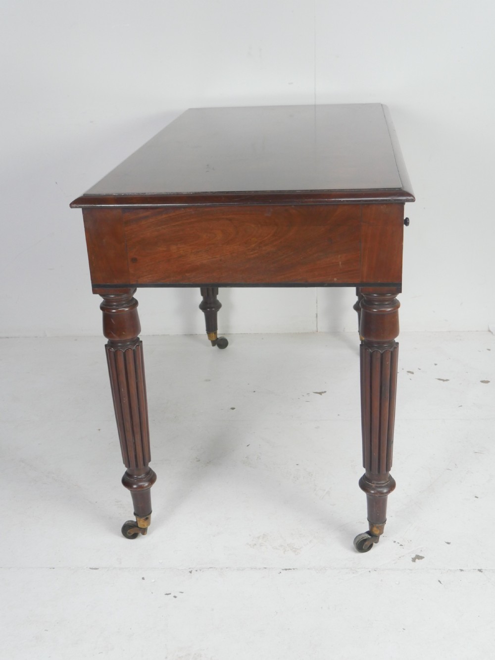 An English Regency mahogany freestanding writing desk featuring three frieze drawers with small turned knobs, supported by four reeded legs on brass castors. View X