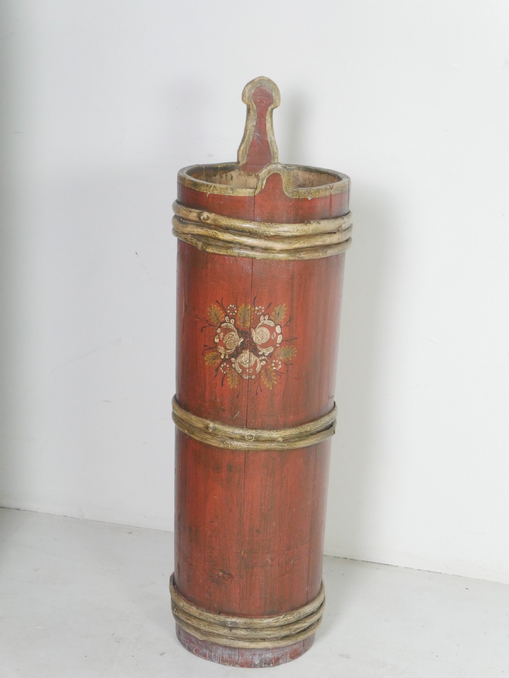 A tall, cylindrical red-painted wooden umbrella stand featuring hand-painted floral motifs and raised wooden banding.