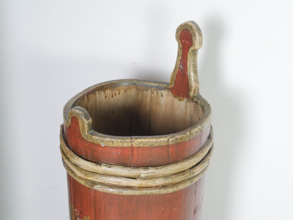 A tall, cylindrical red-painted wooden umbrella stand featuring hand-painted floral motifs and raised wooden banding. View X