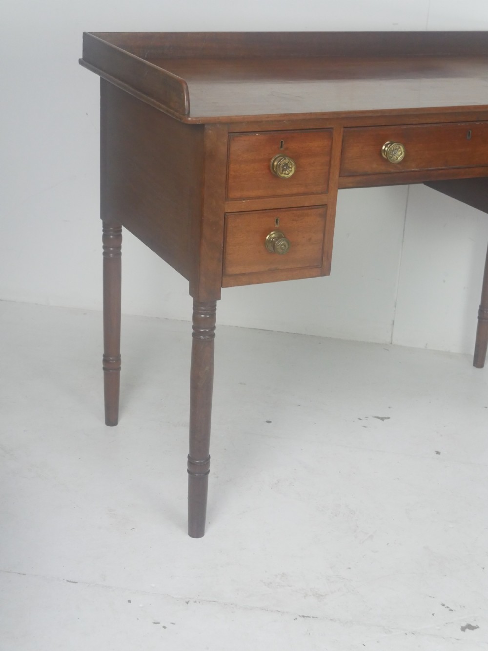 A Regency period mahogany writing desk featuring a low rear gallery, five drawers with brass handles, and four turned legs. View IV
