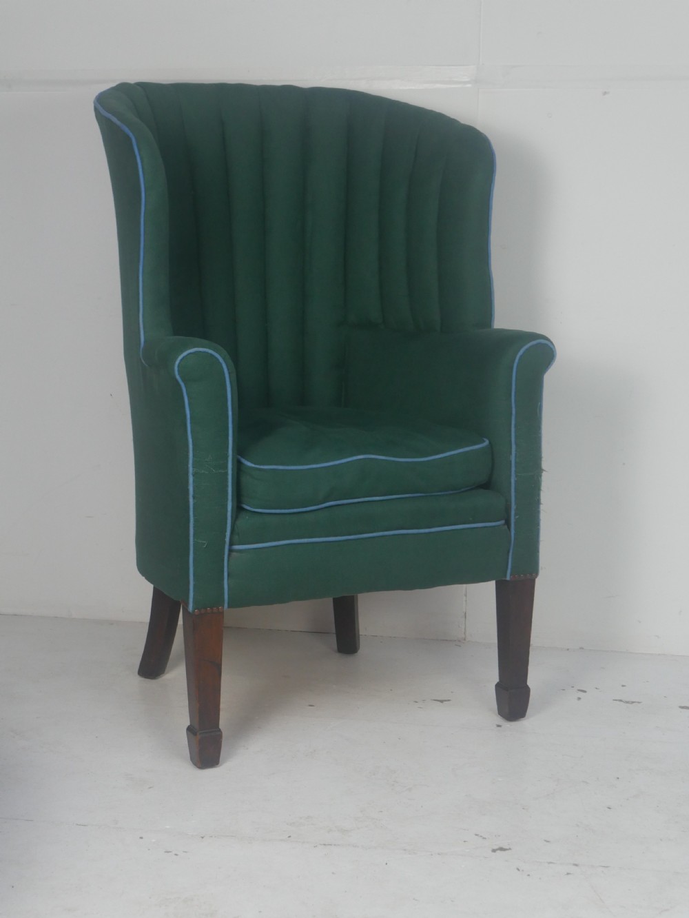 A forest green upholstered barrel back armchair with vertical channel tufting, light blue contrast piping, and dark tapered square legs.