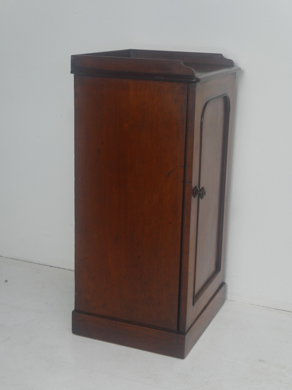 A 19th-century mahogany bedside cupboard featuring a single arched panel door, turned wooden knob, top galley edge, and a solid plinth base. View IV