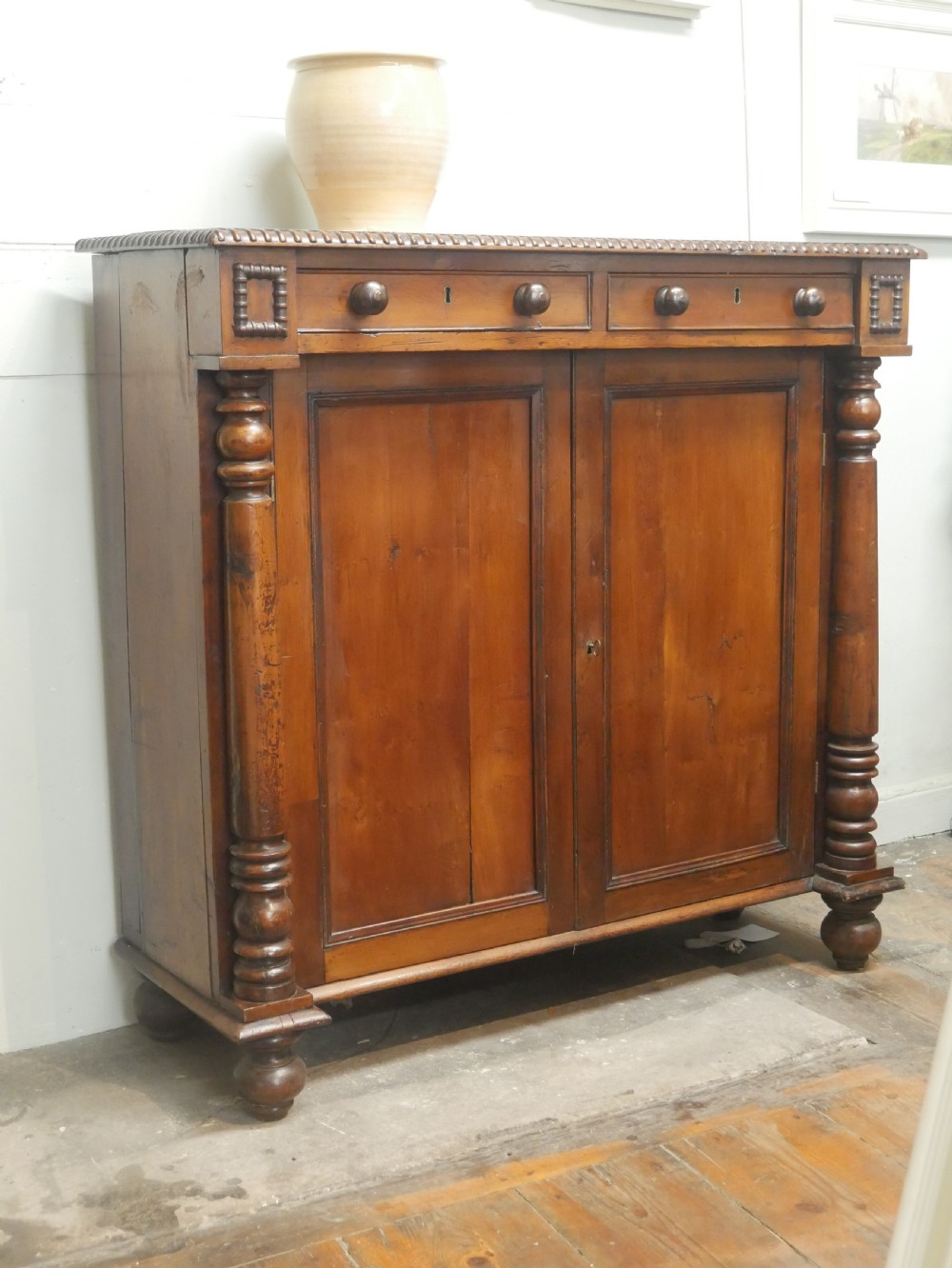 A warm brown cedar cupboard featuring two top drawers, twin panelled doors, and carved side columns supported by turned feet.