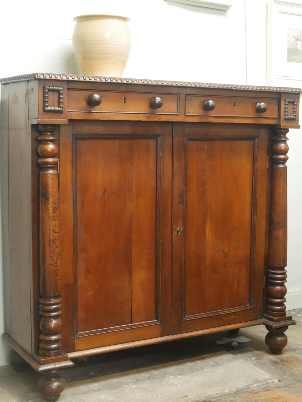 A warm brown cedar cupboard featuring two top drawers, twin panelled doors, and carved side columns supported by turned feet. View XIV