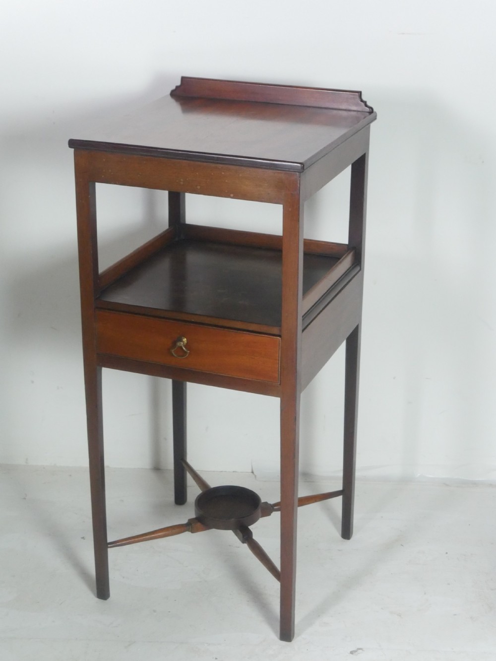 A narrow dark mahogany bedside table featuring a top surface with a gallery back, an open central shelf, a single drawer with a brass drop handle, and cross stretchers with a turned dish centre.