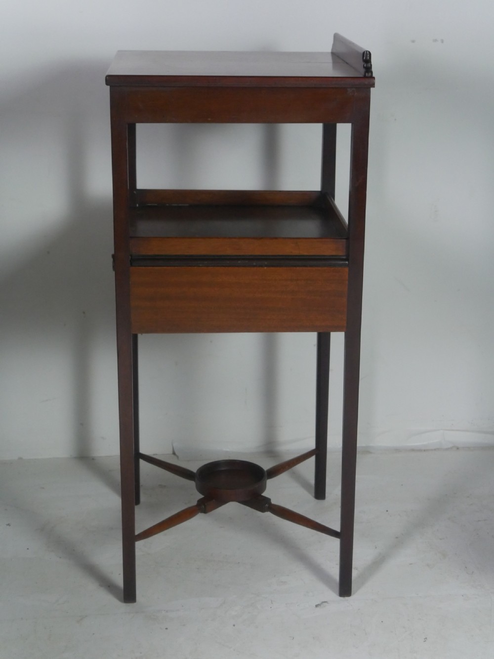A narrow dark mahogany bedside table featuring a top surface with a gallery back, an open central shelf, a single drawer with a brass drop handle, and cross stretchers with a turned dish centre. View V