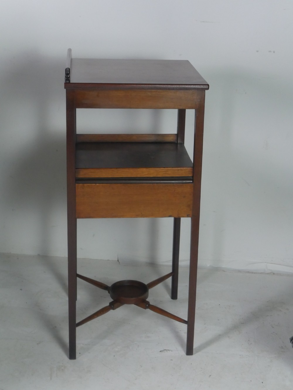 A narrow dark mahogany bedside table featuring a top surface with a gallery back, an open central shelf, a single drawer with a brass drop handle, and cross stretchers with a turned dish centre. View VI