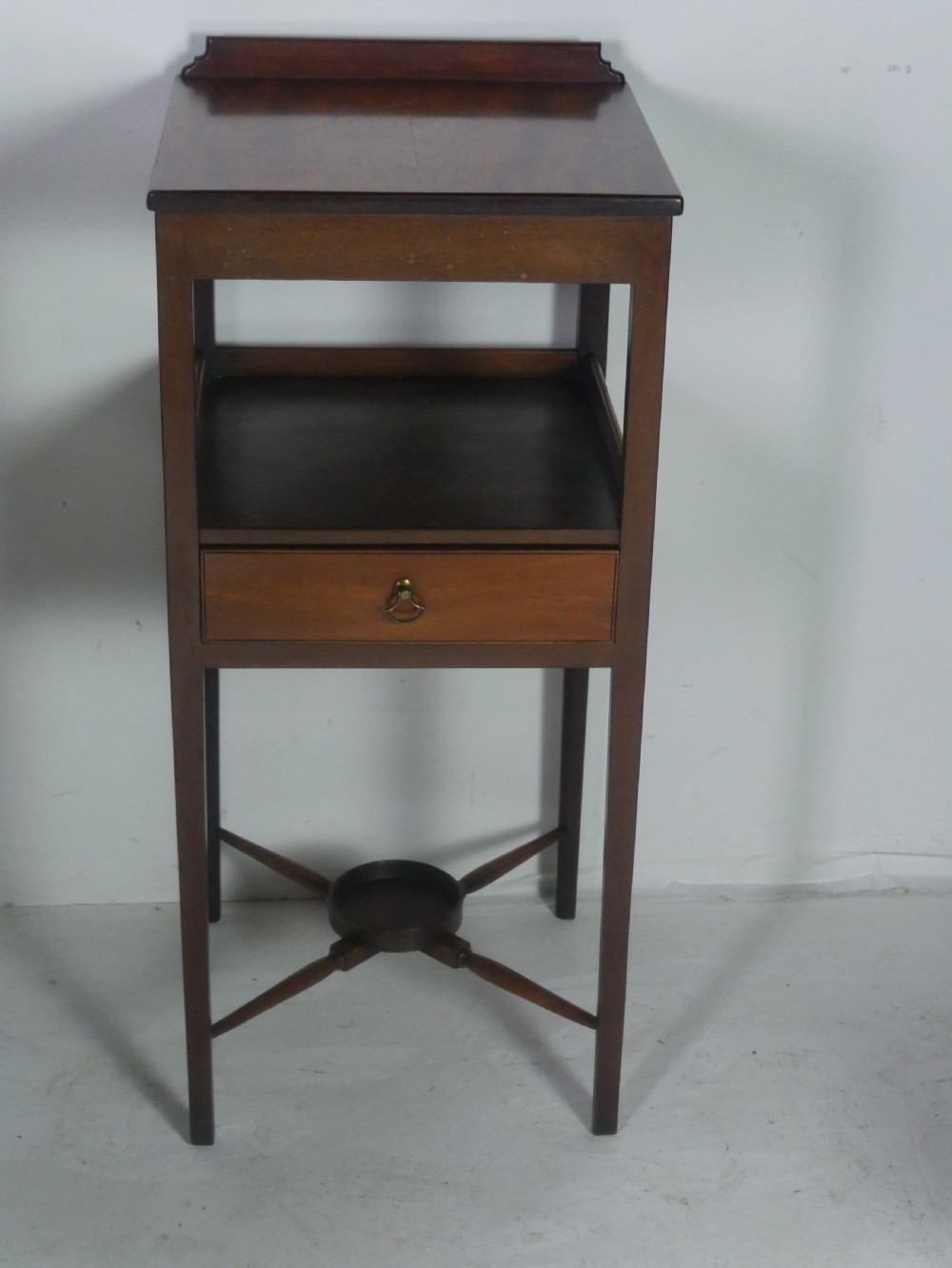 A narrow dark mahogany bedside table featuring a top surface with a gallery back, an open central shelf, a single drawer with a brass drop handle, and cross stretchers with a turned dish centre. View VII