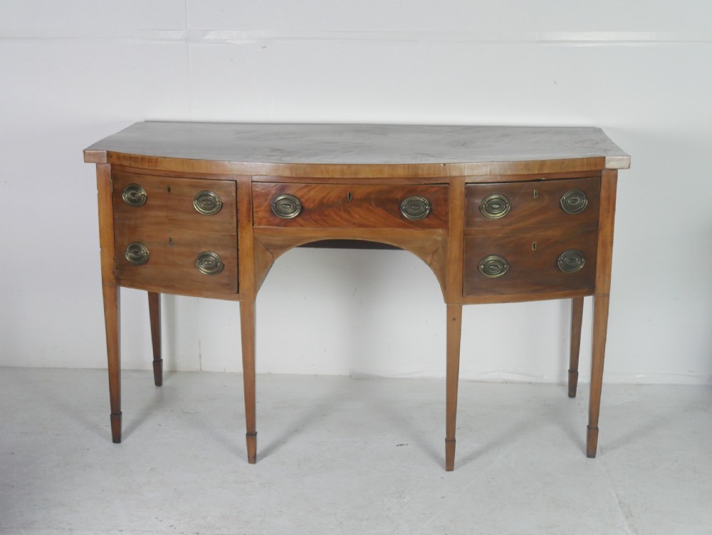 A Georgian mahogany bow-front sideboard featuring a central arch aperture, multiple drawers with oval brass handles, and square tapered legs.