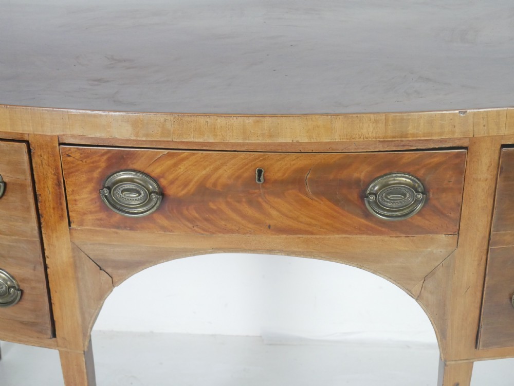 A Georgian mahogany bow-front sideboard featuring a central arch aperture, multiple drawers with oval brass handles, and square tapered legs. View III