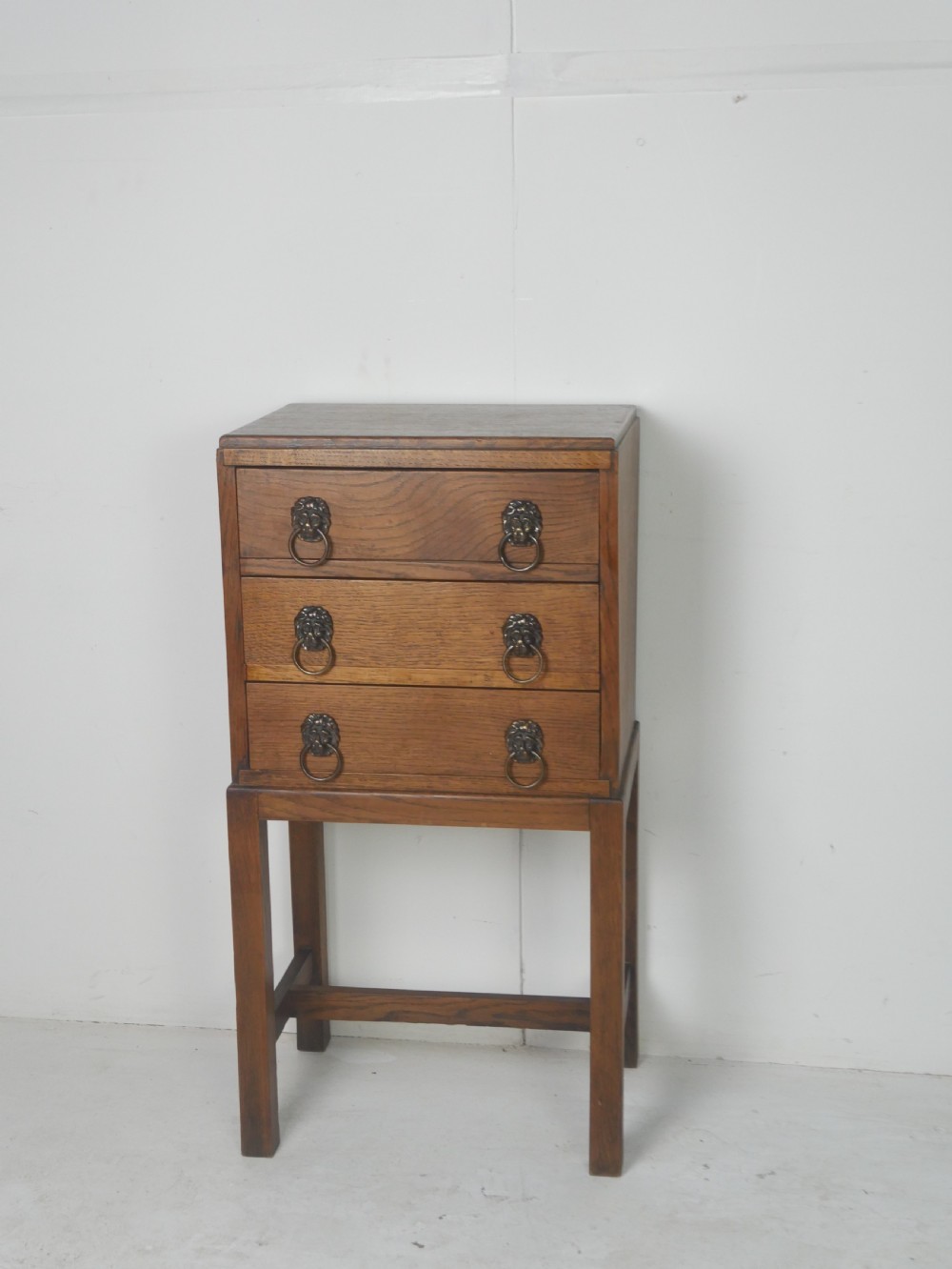 A small medium-brown oak chest featuring three drawers with brass lion mask ring handles, elevated on tall straight legs with an H-stretcher base.