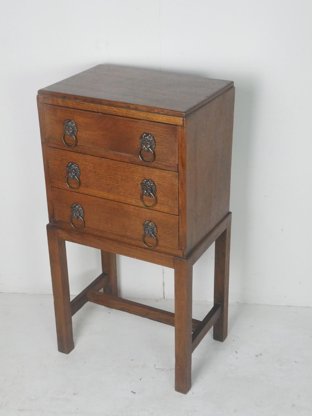 A small medium-brown oak chest featuring three drawers with brass lion mask ring handles, elevated on tall straight legs with an H-stretcher base. View V